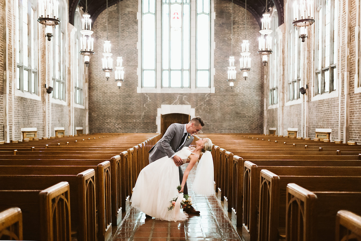 Groom dips his bride in the aisle between wood church pews inside a stone church with large windows all around