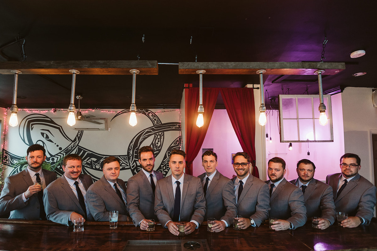 Groom and groomsmen wearing gray suits and dark blue ties line up at wooden bar of The Bitter Alibi
