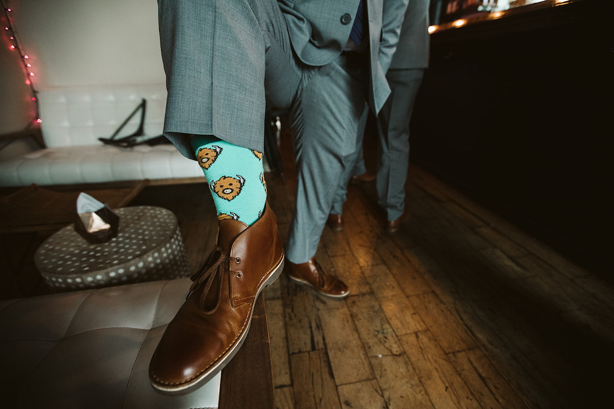 Groomsman pulls up pant leg to show bright blue socks with buffalos on them inside his brown leather ankle boots.