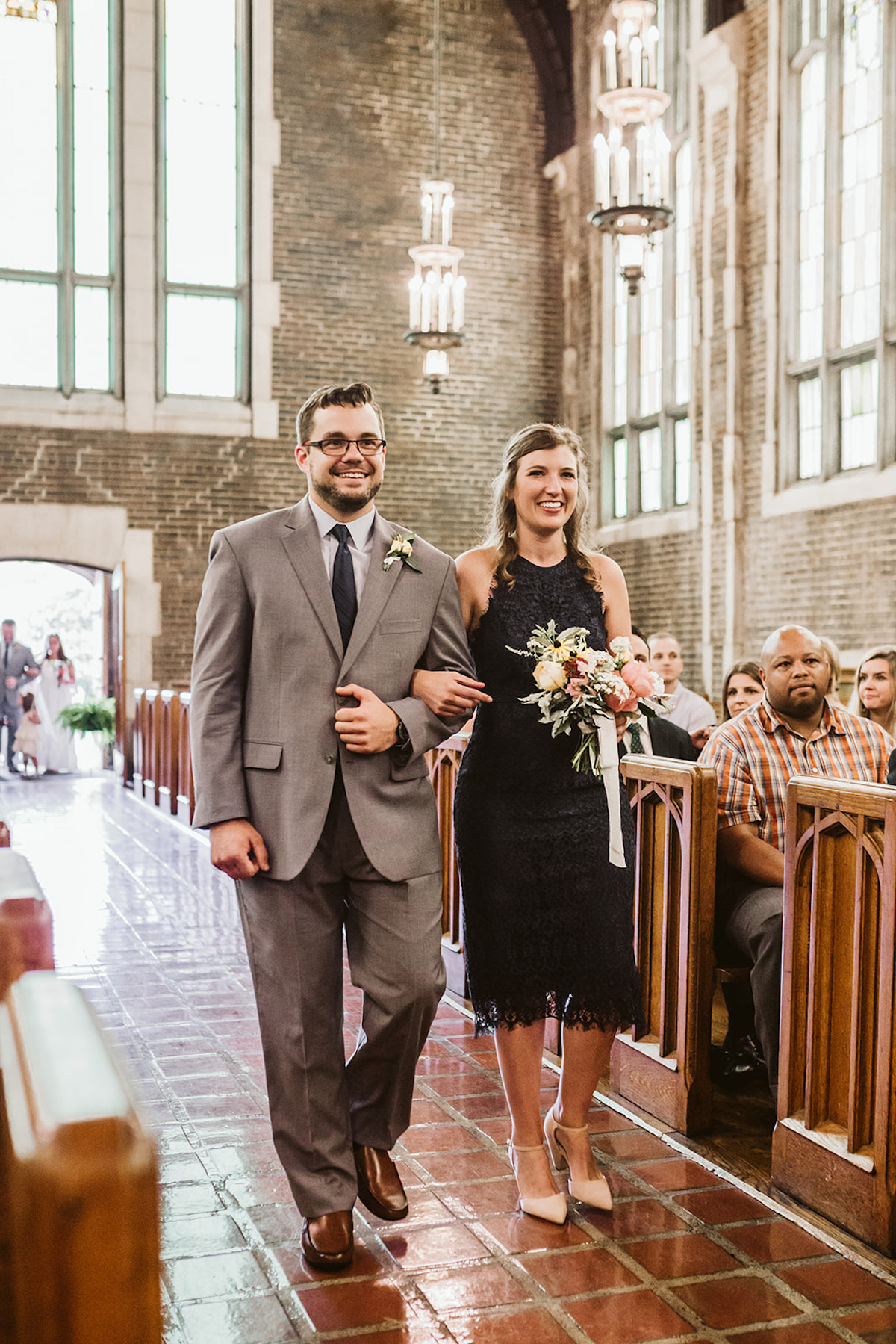 Groomsman and bridesmaid walk brick aisle. Guests sit in wood pews inside high arched ceiling with large windows all around.