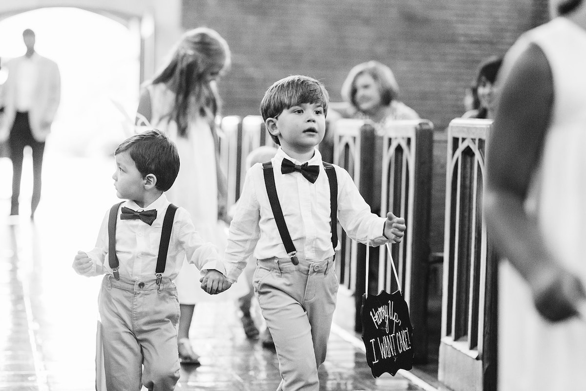 Young ring bearers wearing suspenders and bowties hold hands and walk down the aisle