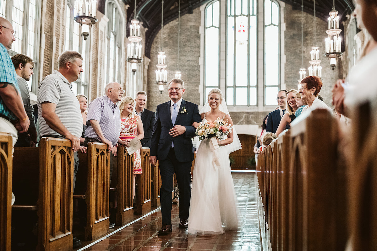 Bride and father walk brick aisle. Guests stand in wood pews inside high arched ceiling with large windows all around.