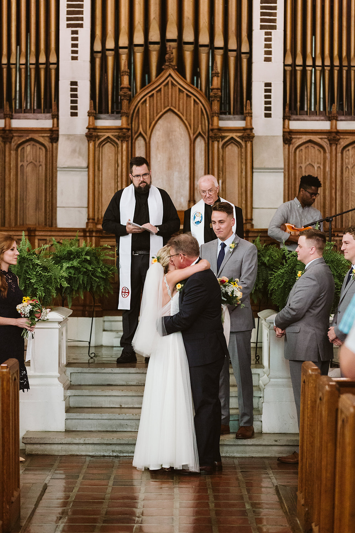 Bride hugs her father while groom and two priests watch