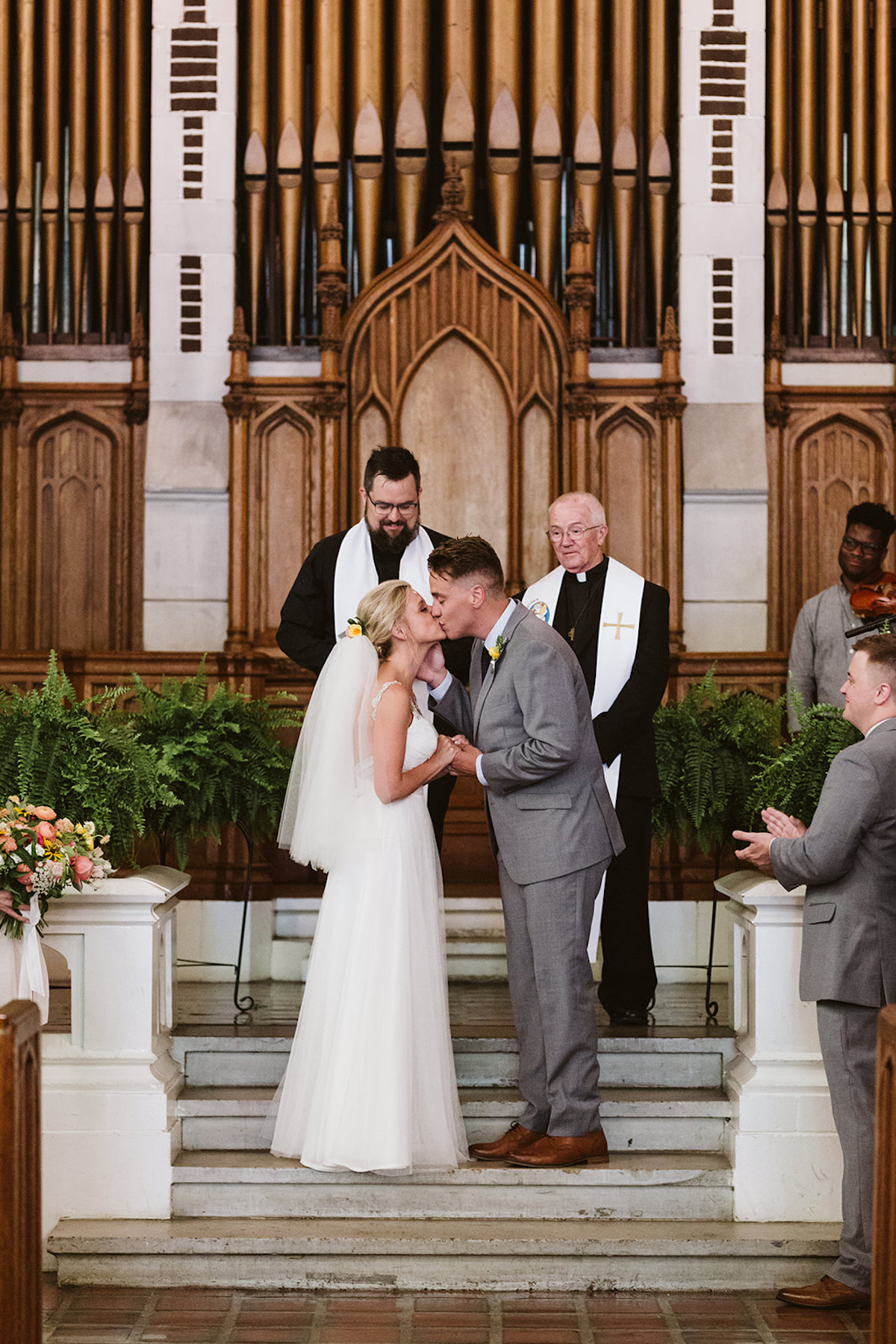 Bride and groom stand on stone steps at the altar, kissing. Two priests and church organ behind them.