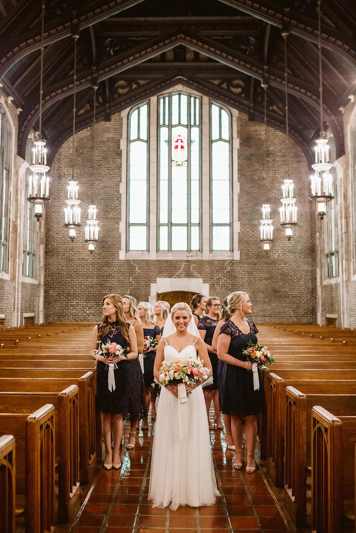 Bride and bridesmaids stand between wooden pews, a large window behind them. She holds a pink, peach, and yellow bouquet.