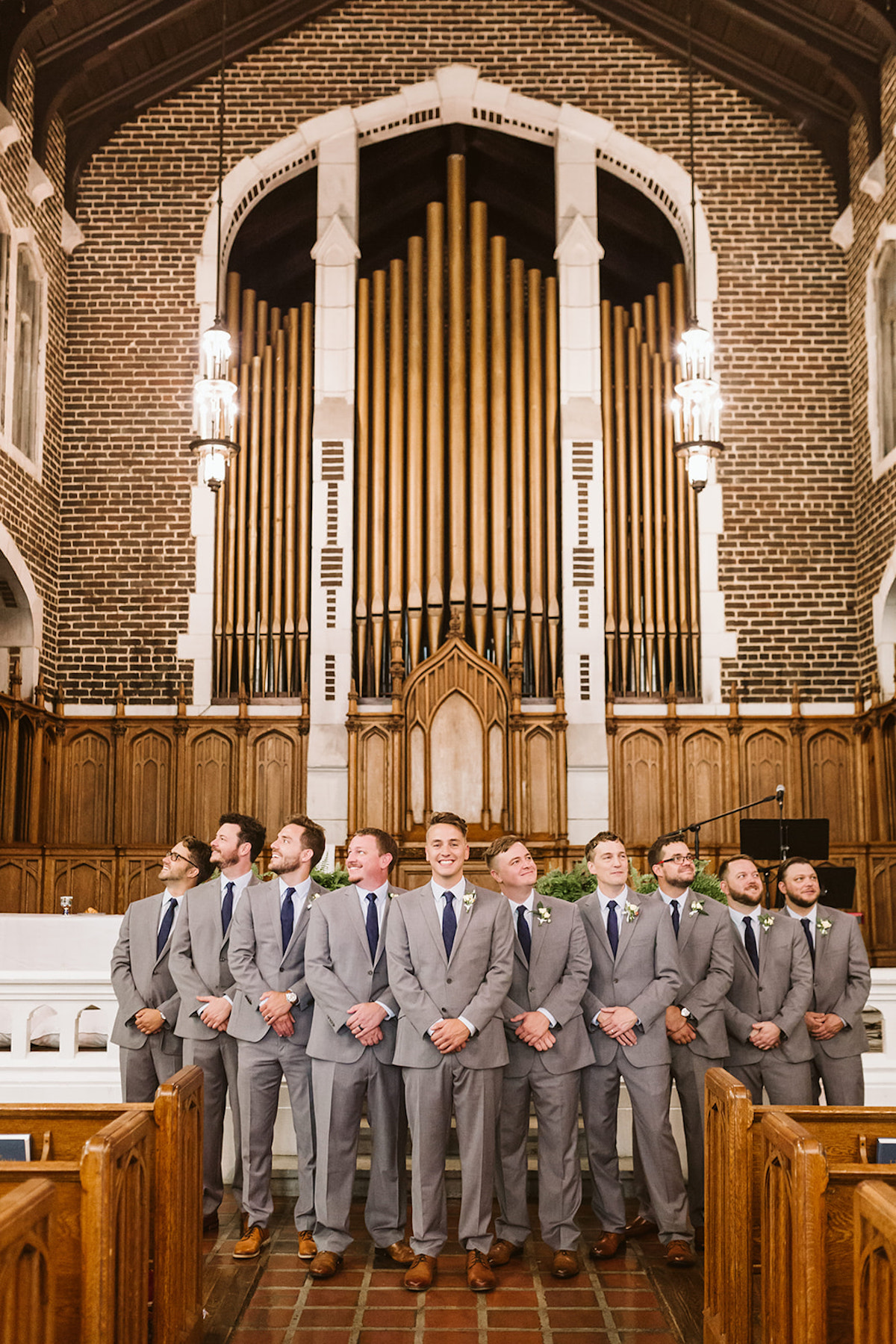 Groom and groomsmen stand between wooden pews in front of large organ pipes in the arched brick sanctuary of Patten Chapel.