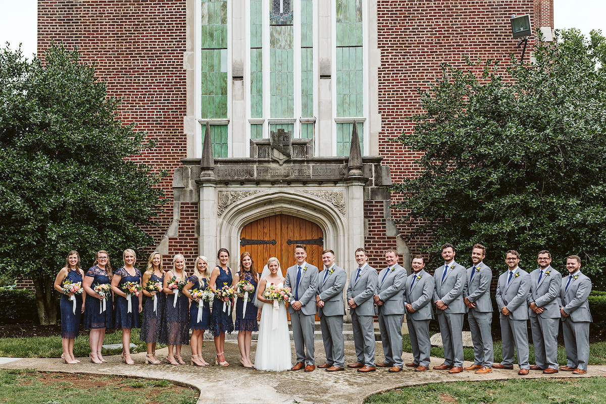 Bridal party stands in front of Patten Chapel's wooden front door, brick exterior, and tall stained glass windows