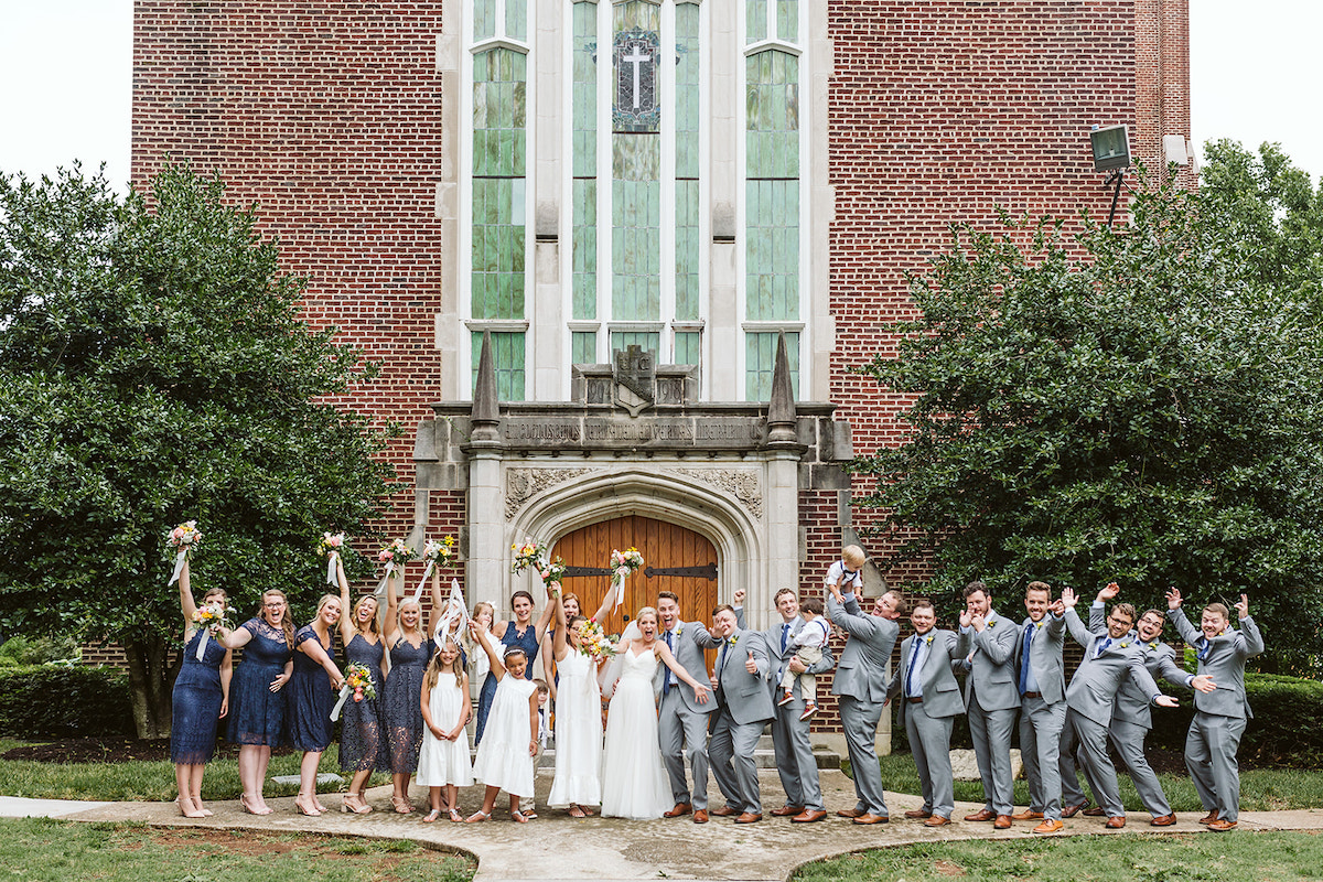 Bridal party stands cheering in front of Patten Chapel's wooden front door, brick exterior, and tall stained glass windows