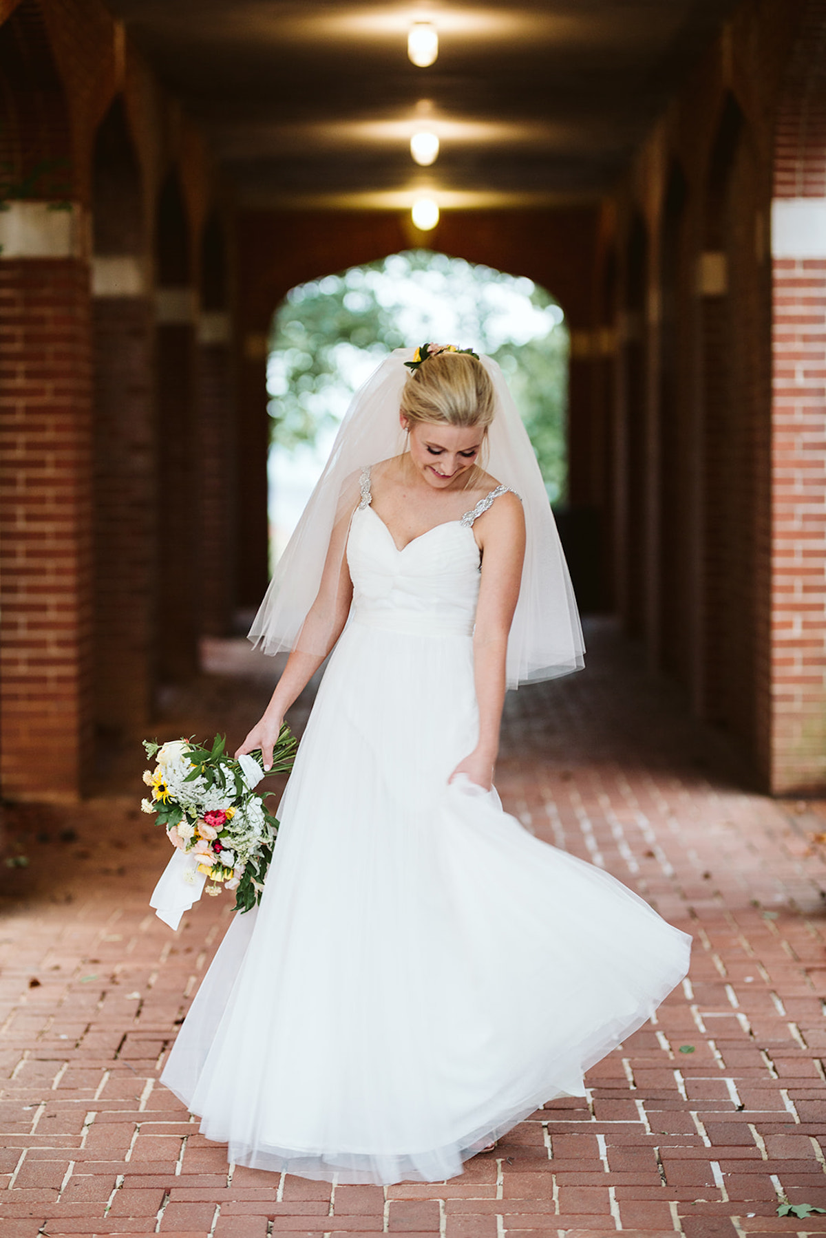 Bride stands in a covered, brick arched walkway holding her bouquet at her side