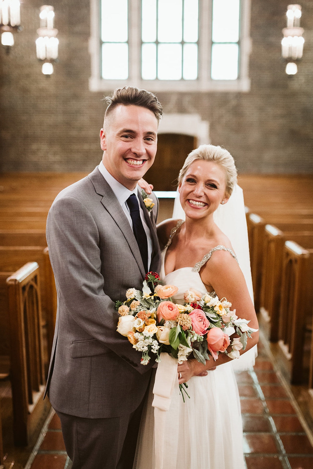 Bride and groom stand between wooden pews, a large window behind them. She holds a colorful pink, peach, and yellow bouquet.