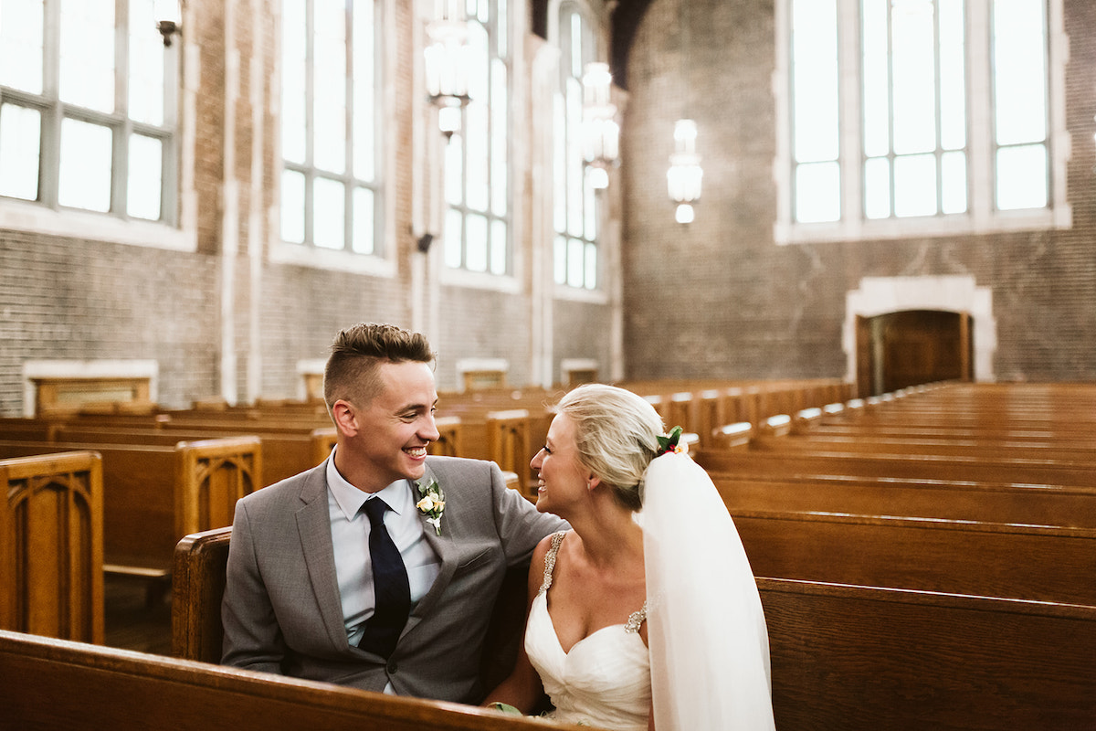 Bride and groom sit smiling at each other in a wooden pew in an arched brick sanctuary, large windows all around the room.
