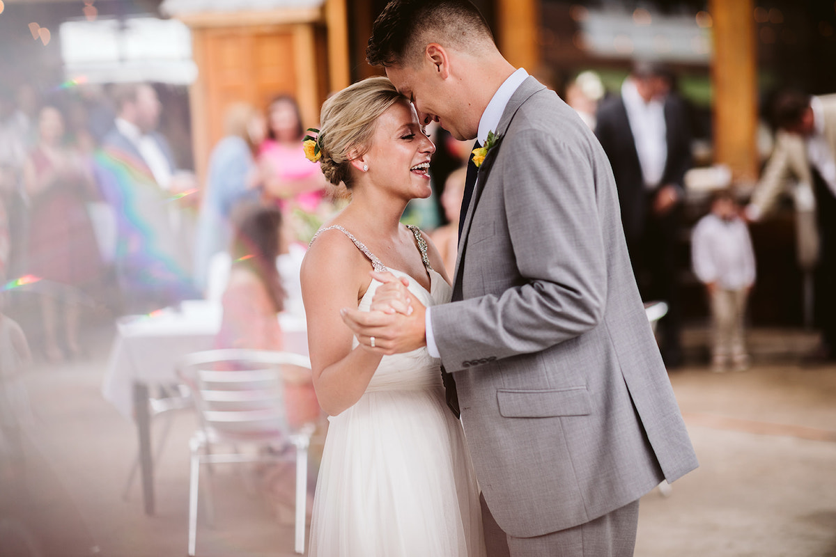 Bride and groom dance, foreheads bowed together. She has yellow flowers in her hair, and he wears a yellow flower boutonniere