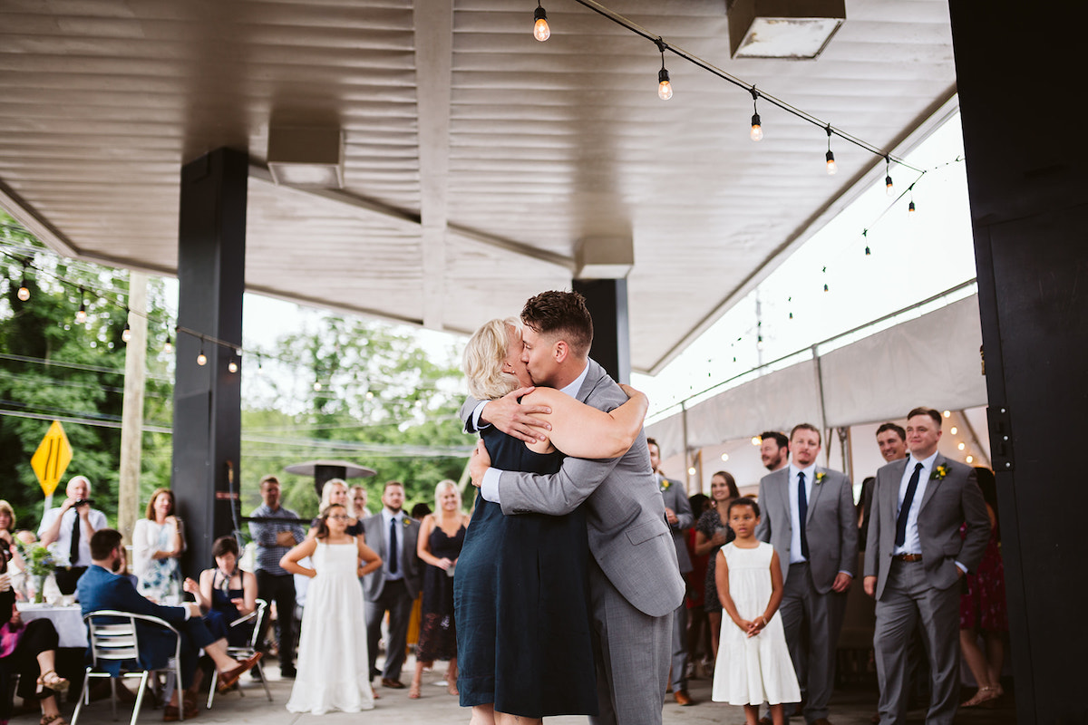 Groom hugs his mother and kisses her cheek under patio cover at The Daily Ration as guests watch.