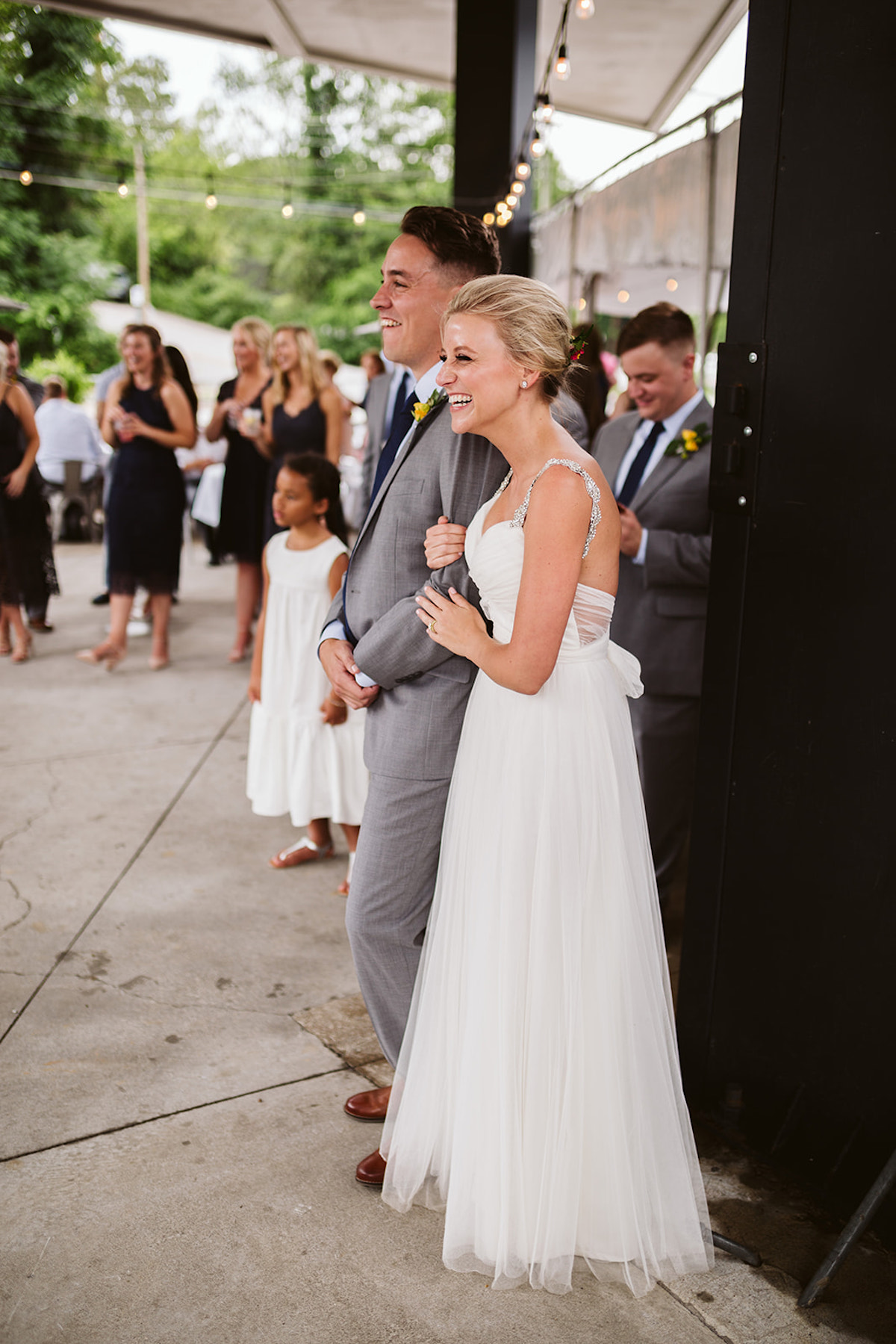 Bride and groom stand with wedding guests under patio cover, laughing