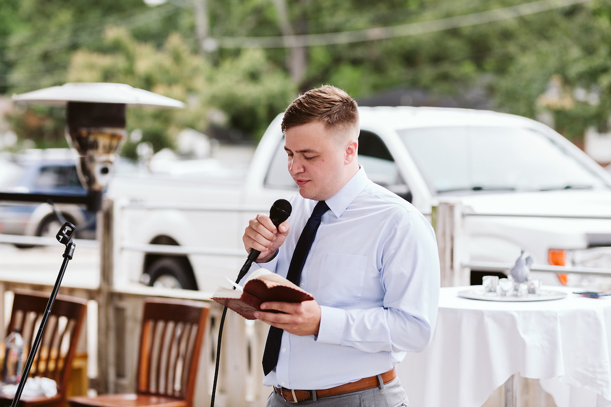 Man wearing light blue shirt and dark blue tie holds microphone and reads his toast from a leather book