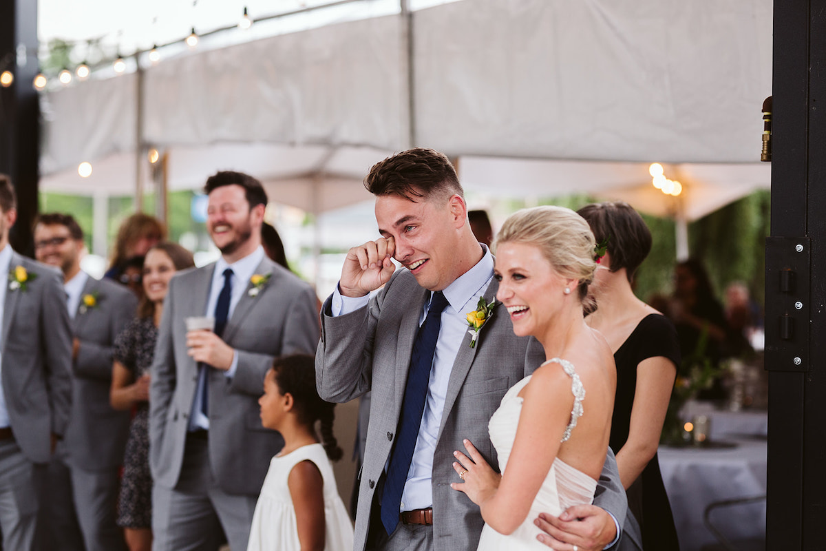 Groom wipes tear from eye while he, bride, and wedding guests laugh