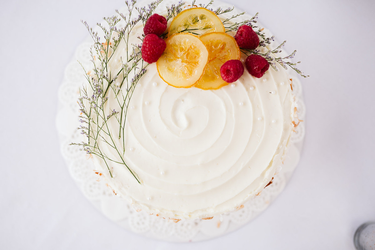 Wedding cake with swirled white frosting topped with a few sprigs of greenery, a few raspberries and slices of candied orange