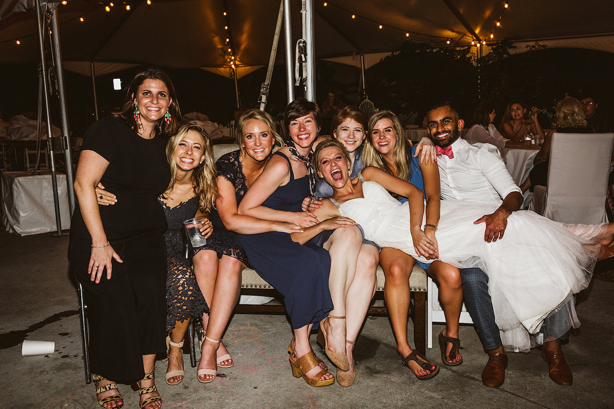 Bride lays across laps of friends under white tent with string patio lights