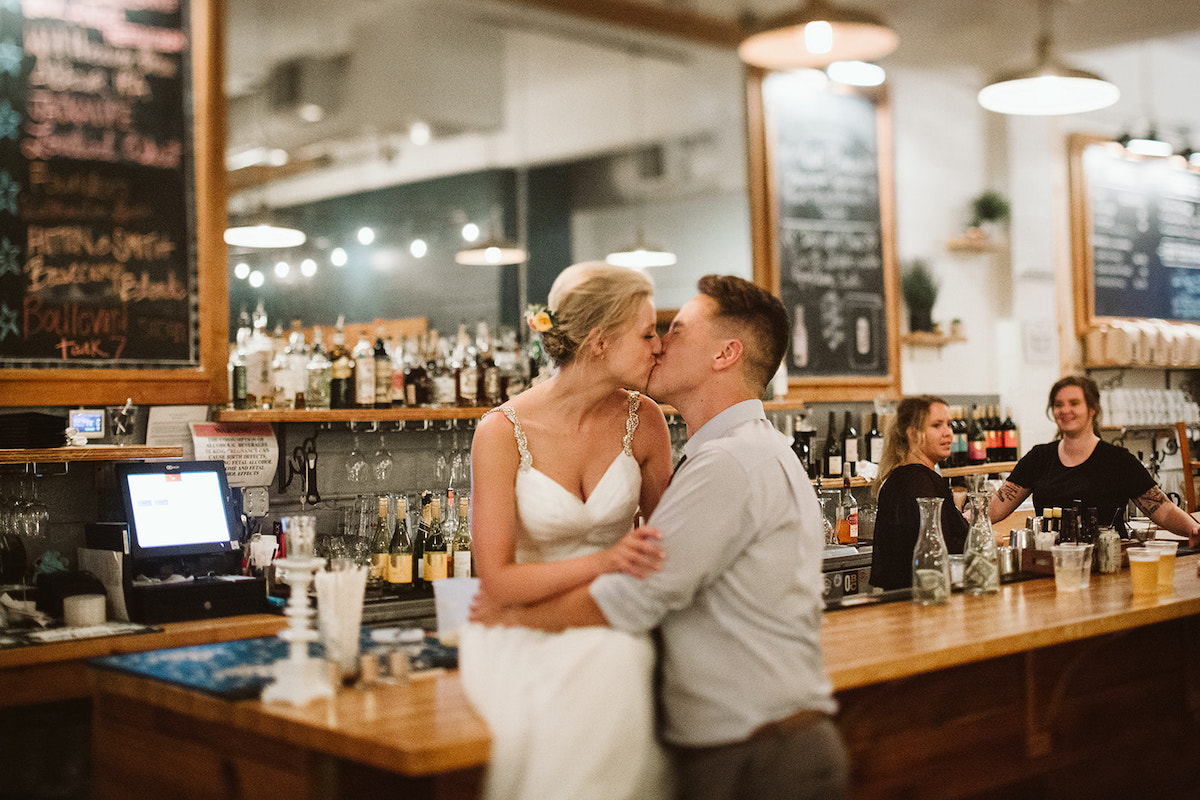 Bride sits on wooden bar while groom kisses her. Bar staff and wine bottles and large mirror are behind them.