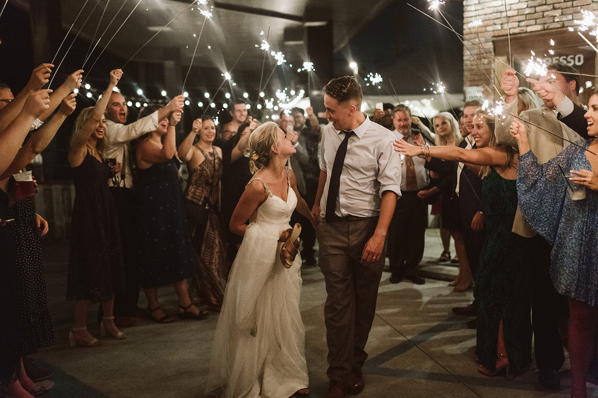 Groom and bride walk through crowd under sparkler tunnel, smiling at each other while guests cheer.