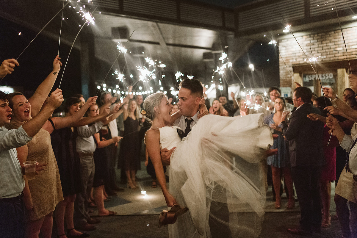 Groom holds bride under sparkler tunnel. They look at each other, cheering.