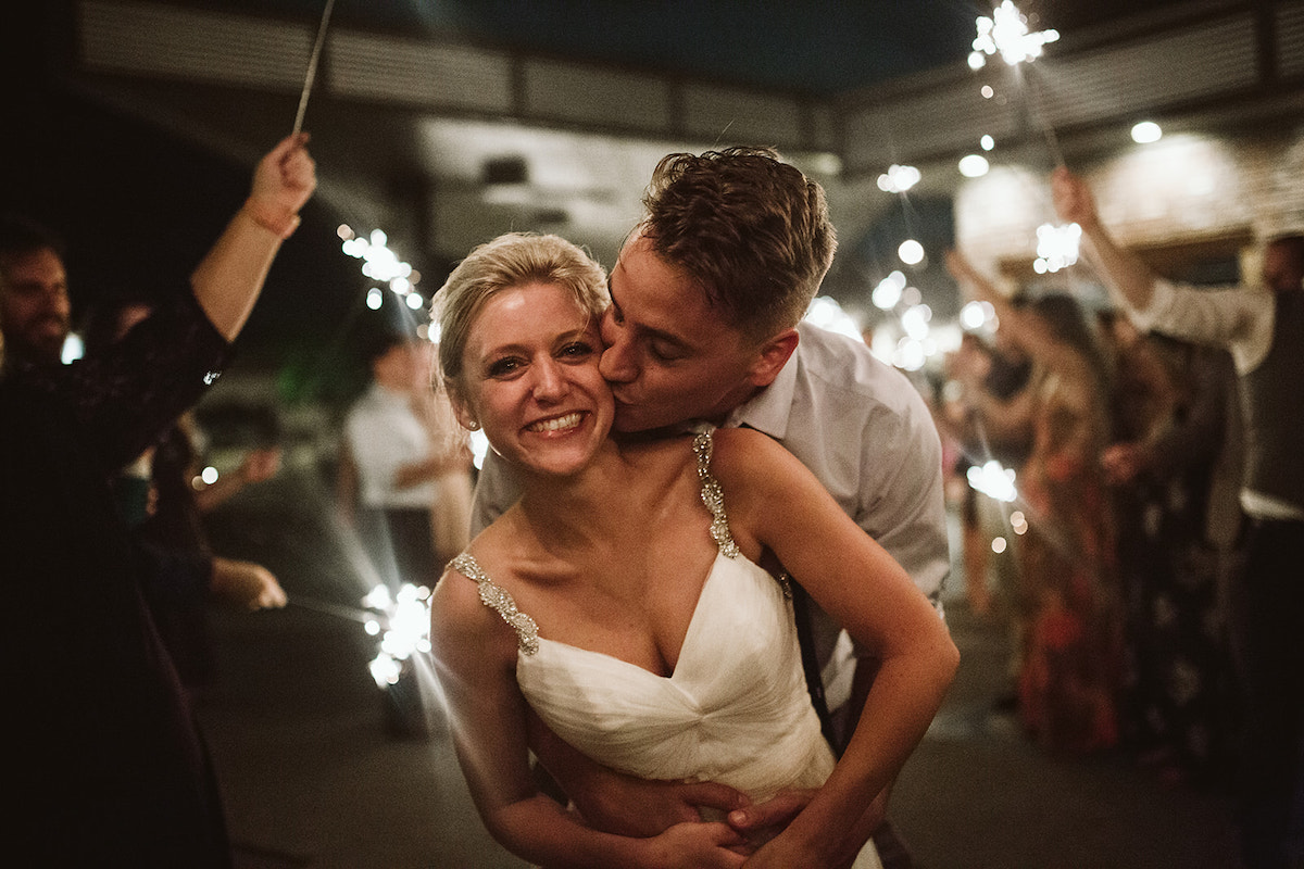 Groom stands behind bride, hugging around her waist and kissing her cheek under sparkler tunnel