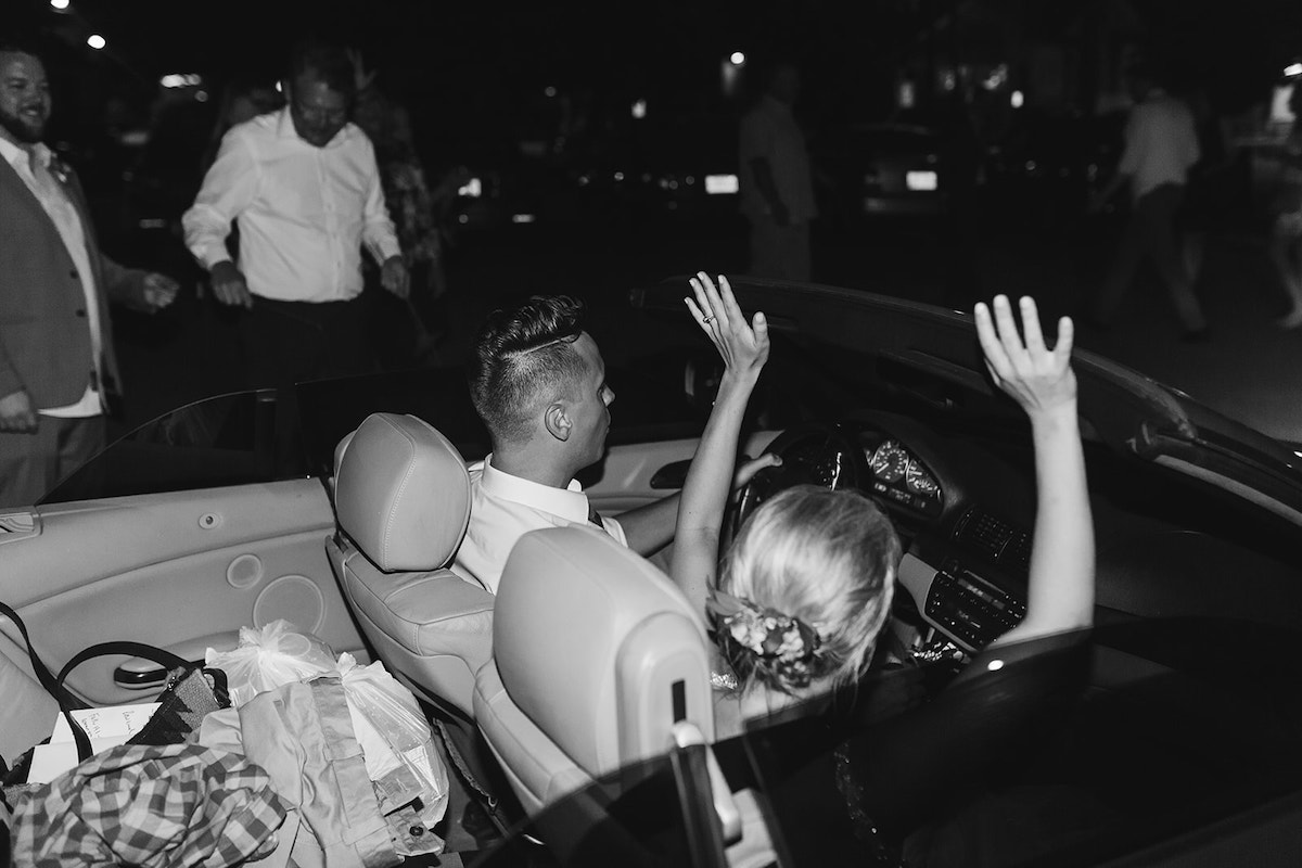 Bride and groom exit in convertible with the roof down, bride's hands raised in the sky