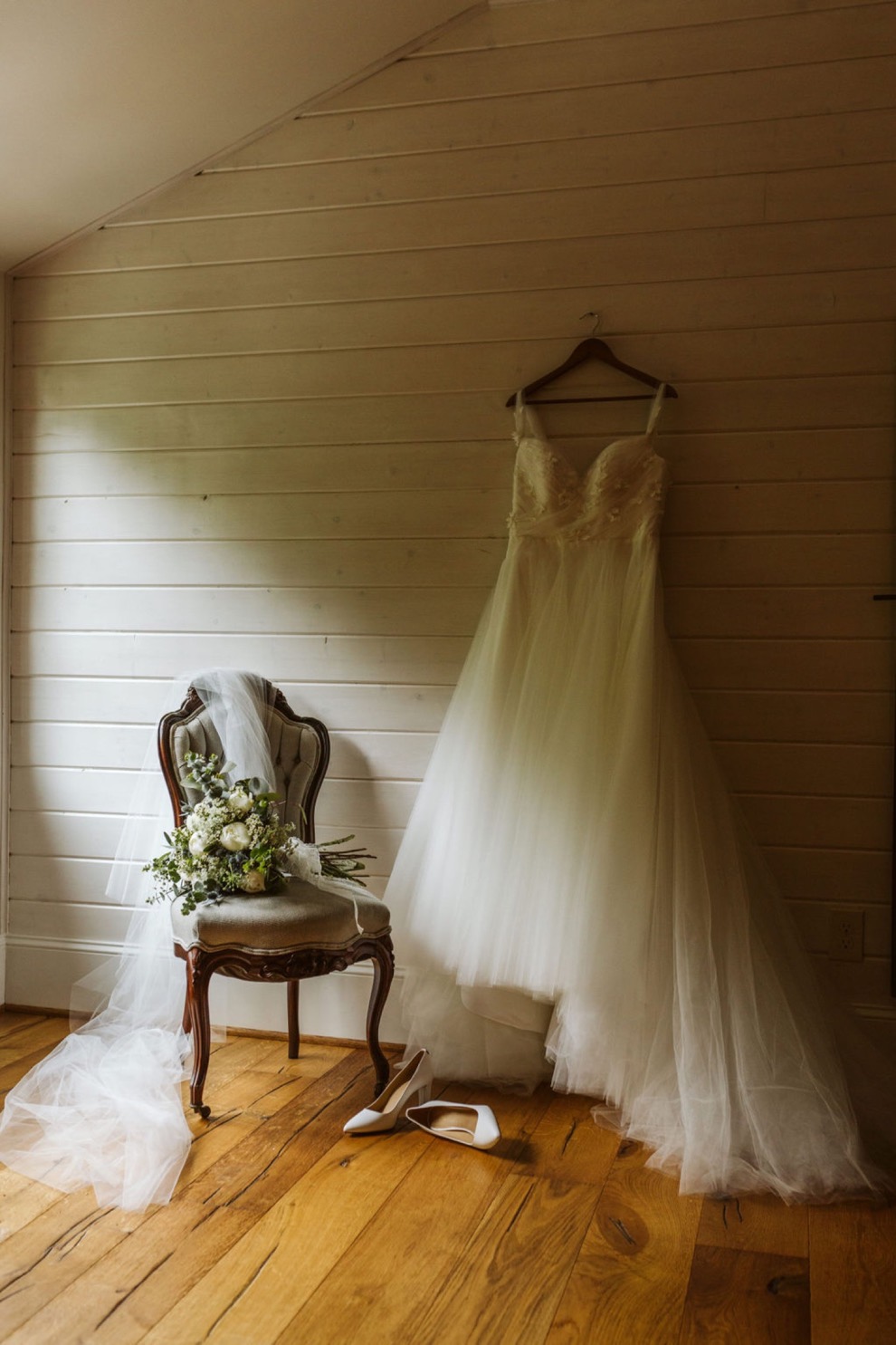 wedding dress hanging on shiplap white wall. Bride's veil and bouquet sit on an antique chair.