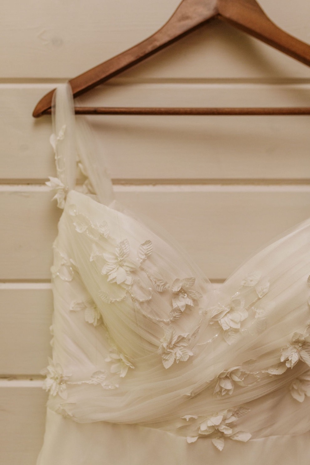 lace appliqué wedding dress hangs on a wood hanger on a white, shiplap wall