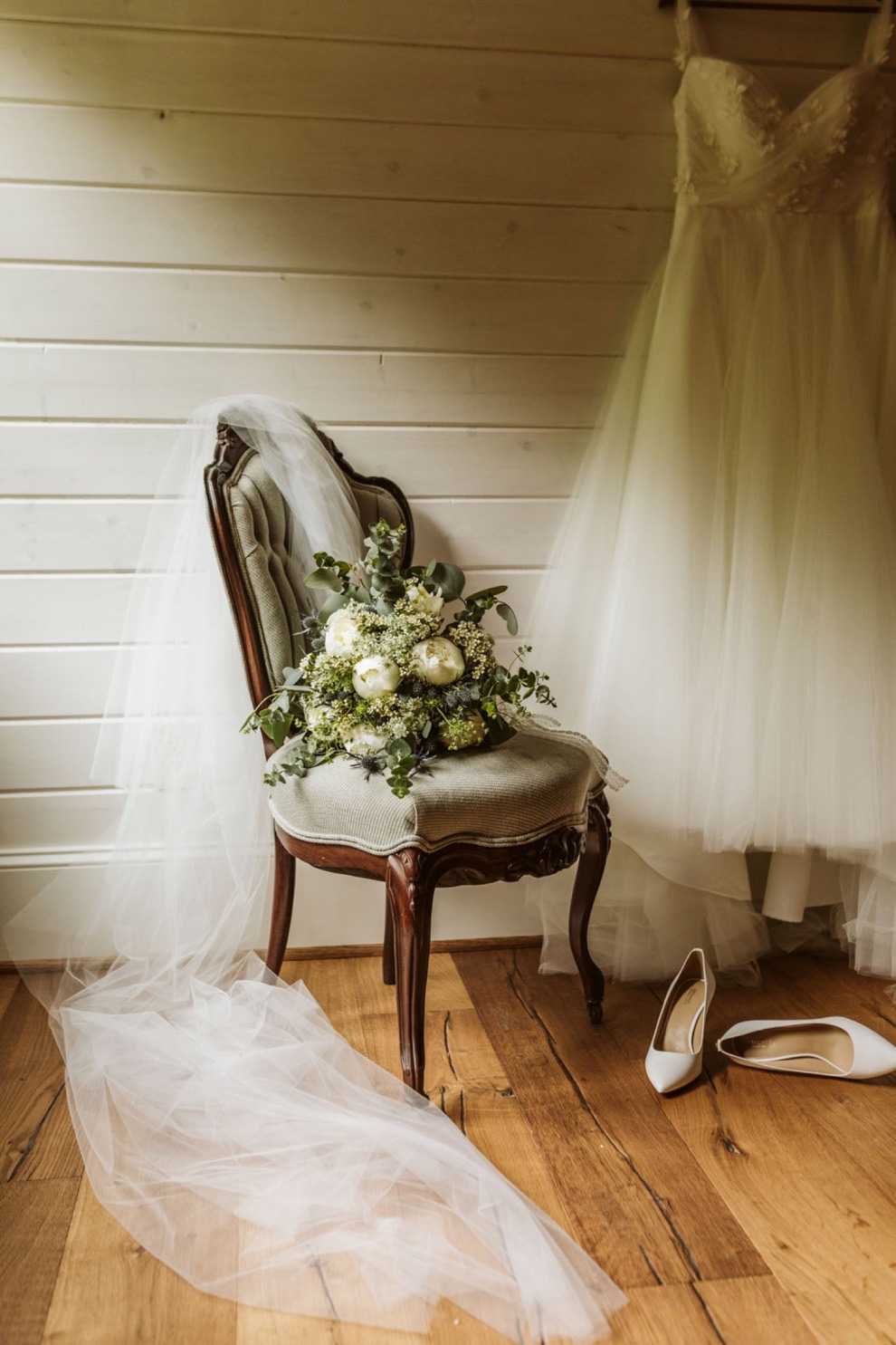 Wedding dress hangs on a shiplap wall with veil and bouquet sitting on an antique chair. White shoes lie on wood plank floor.