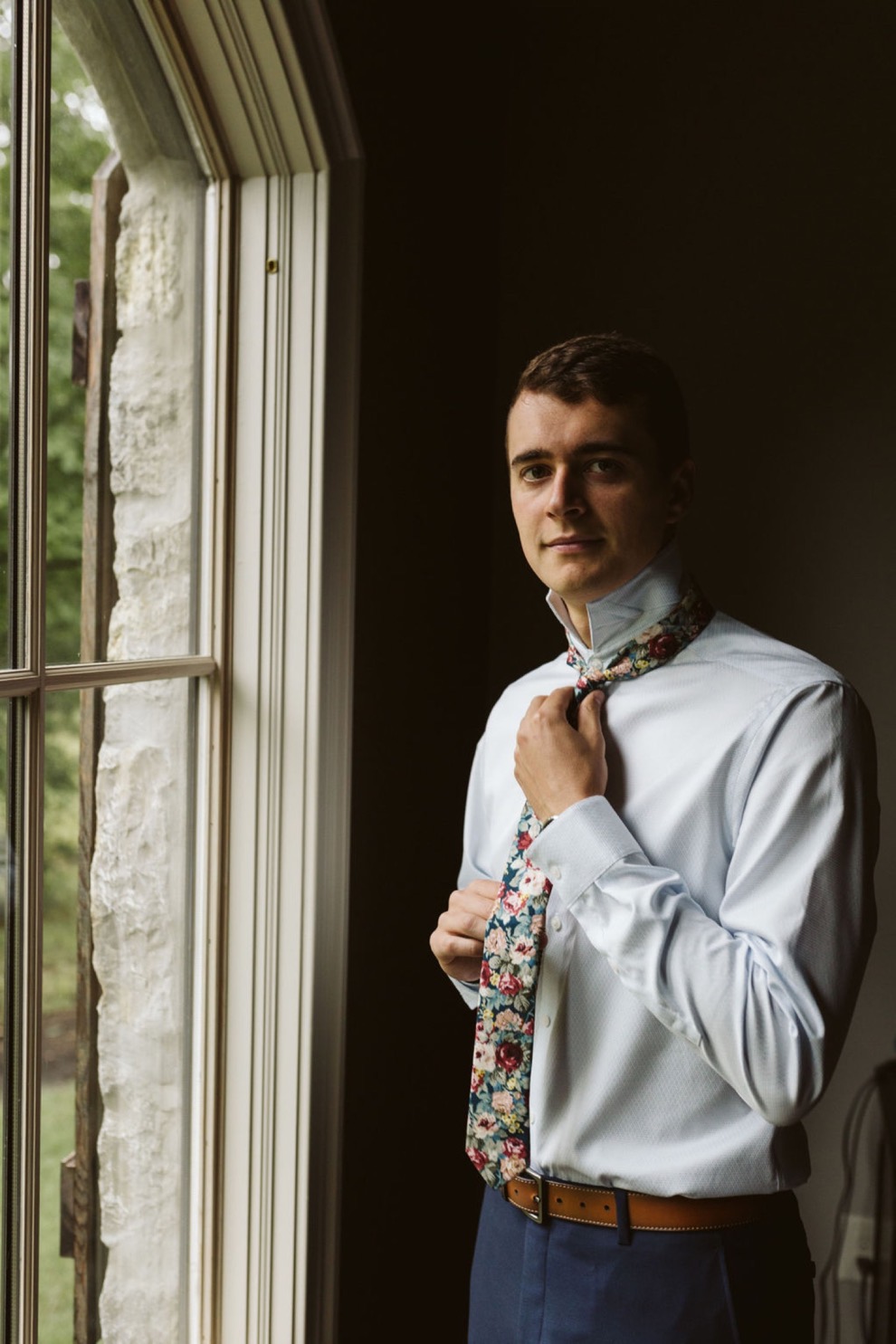 groom in dark blue suit pants and light blue shirt adjusts the knot of his floral tie