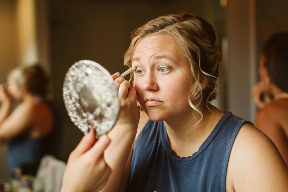 bride putting on eye shadow while a friend holds up a round silver mirror for her
