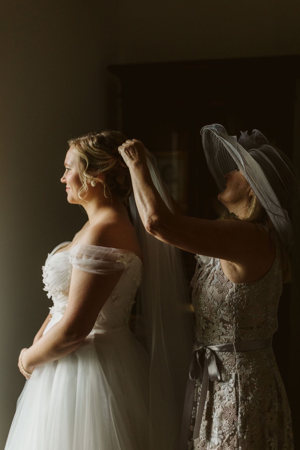 bride's mother tucking veil into her dauther's hair. Bride's mother wears a wide gray hat.