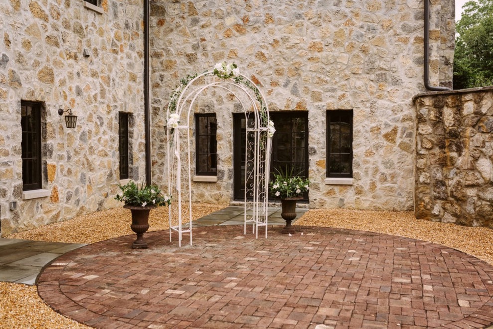 backyard wedding ceremony in a wide, stone courtyard. A white metal wedding arch framed by two large potted plants.
