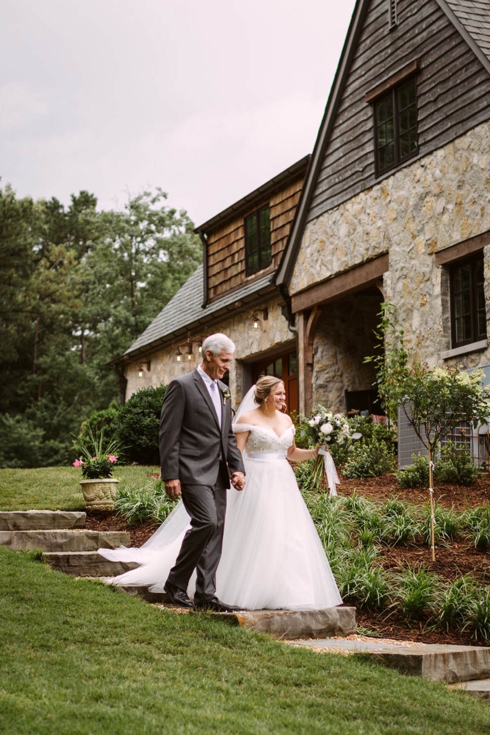 bride's father escorts her down wide stone steps around the side of a stone house