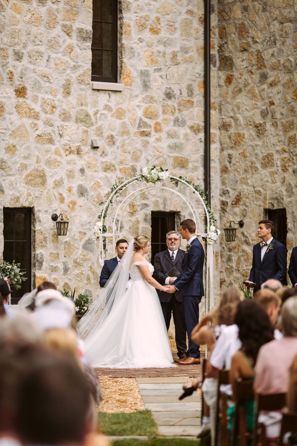 bride and groom holding hands under simple white metal wedding arch in front of a stone house