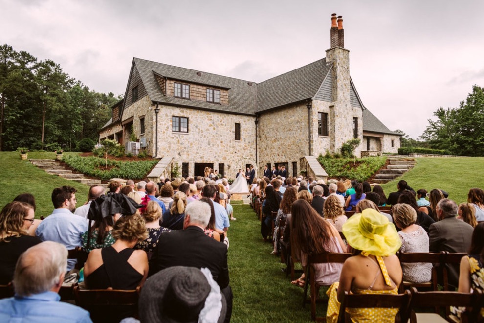 wedding guests sit in wooden folding chairs behind a large, stone house for backyard wedding in Calhoun, Georgia