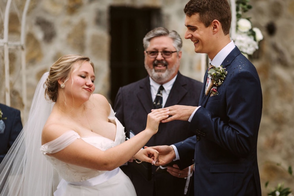 bride sliding ring onto groom's finger while making a funny face. He laughs.