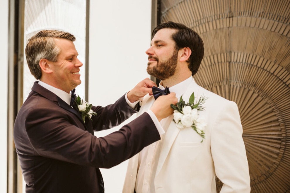 man helping groom in white jacket fix his bowtie