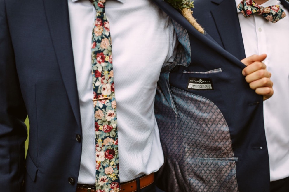 groom holds open blue suit jacket to show his narrow floral tie clipped to his light blue shirt