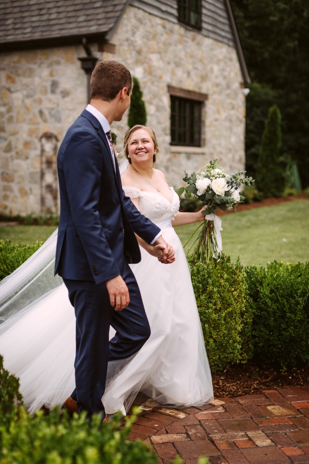 bride and groom smiling at each other as they walk down a brick path between low shrubs