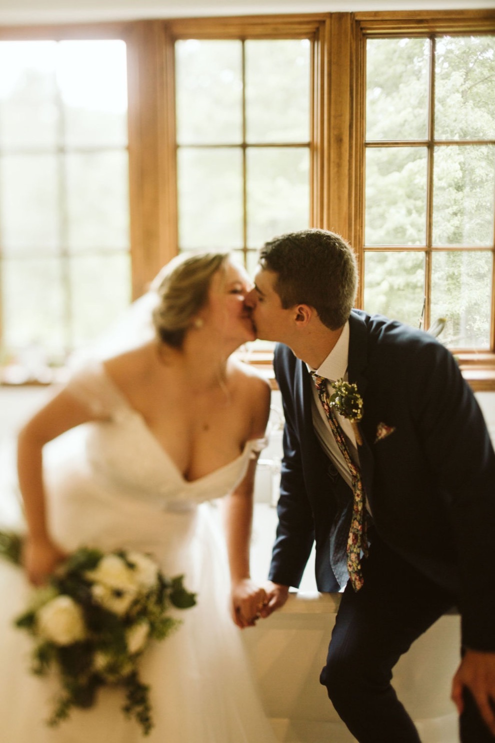 bride and groom kissing in front of window while sitting on the edge of a white claw-foot bathtub in a bright bathroom