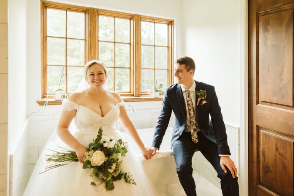 bride and groom holding hands and sitting on the edge of a white claw-foot bathtub in a bright bathroom