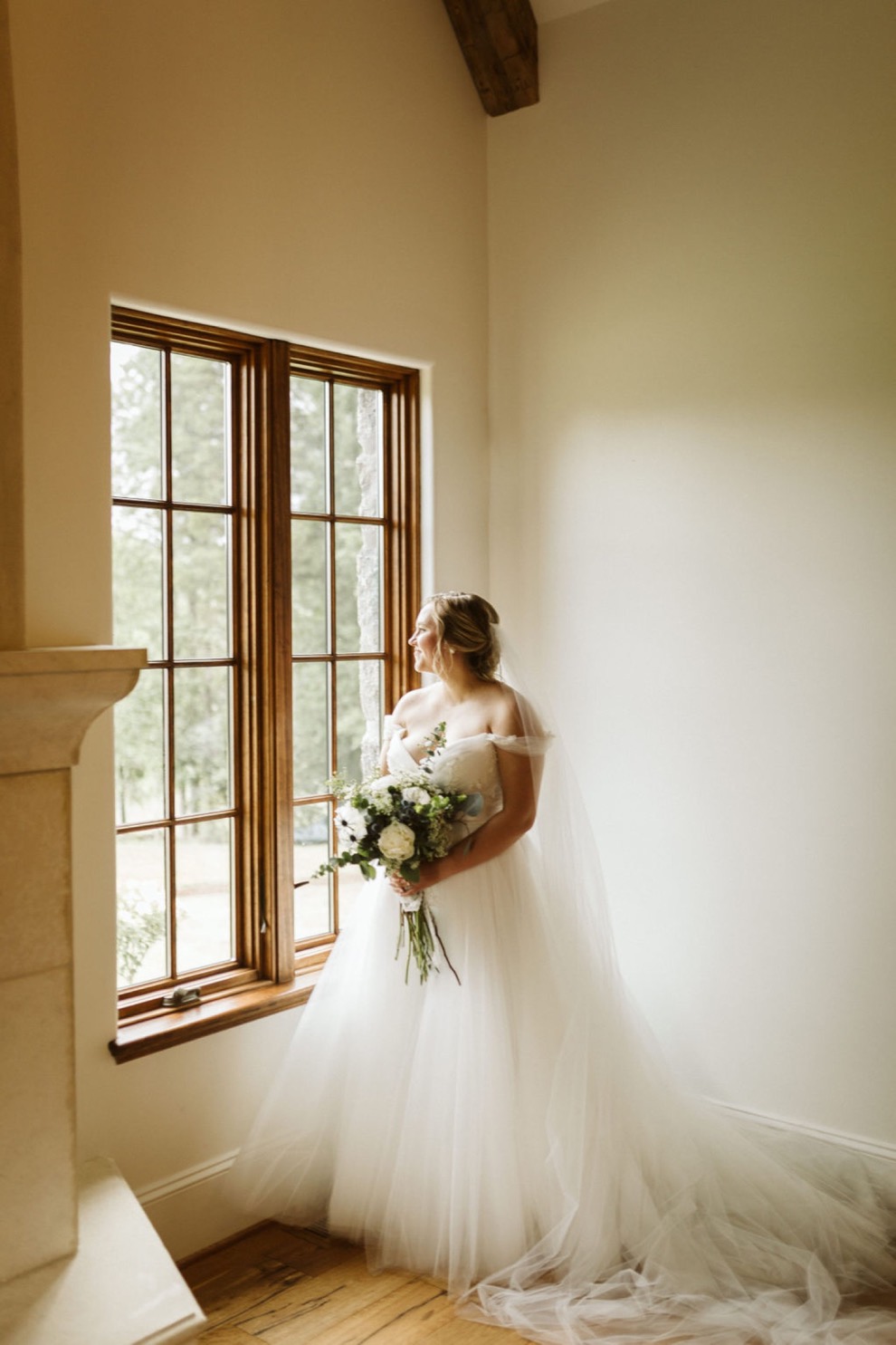 bride standing under a high-peaked ceiling in front of tall window holding a bouquet of large white flowers and greenery