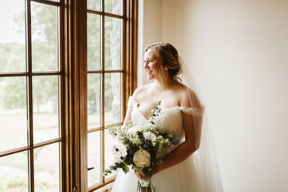 bride standing in front of tall window holding a bouquet of large white flowers and greenery