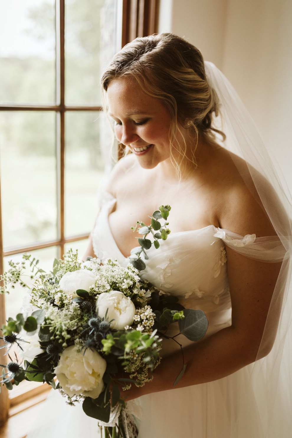 bride standing in front of tall window, smiling down at her bouquet of white flowers and greenery