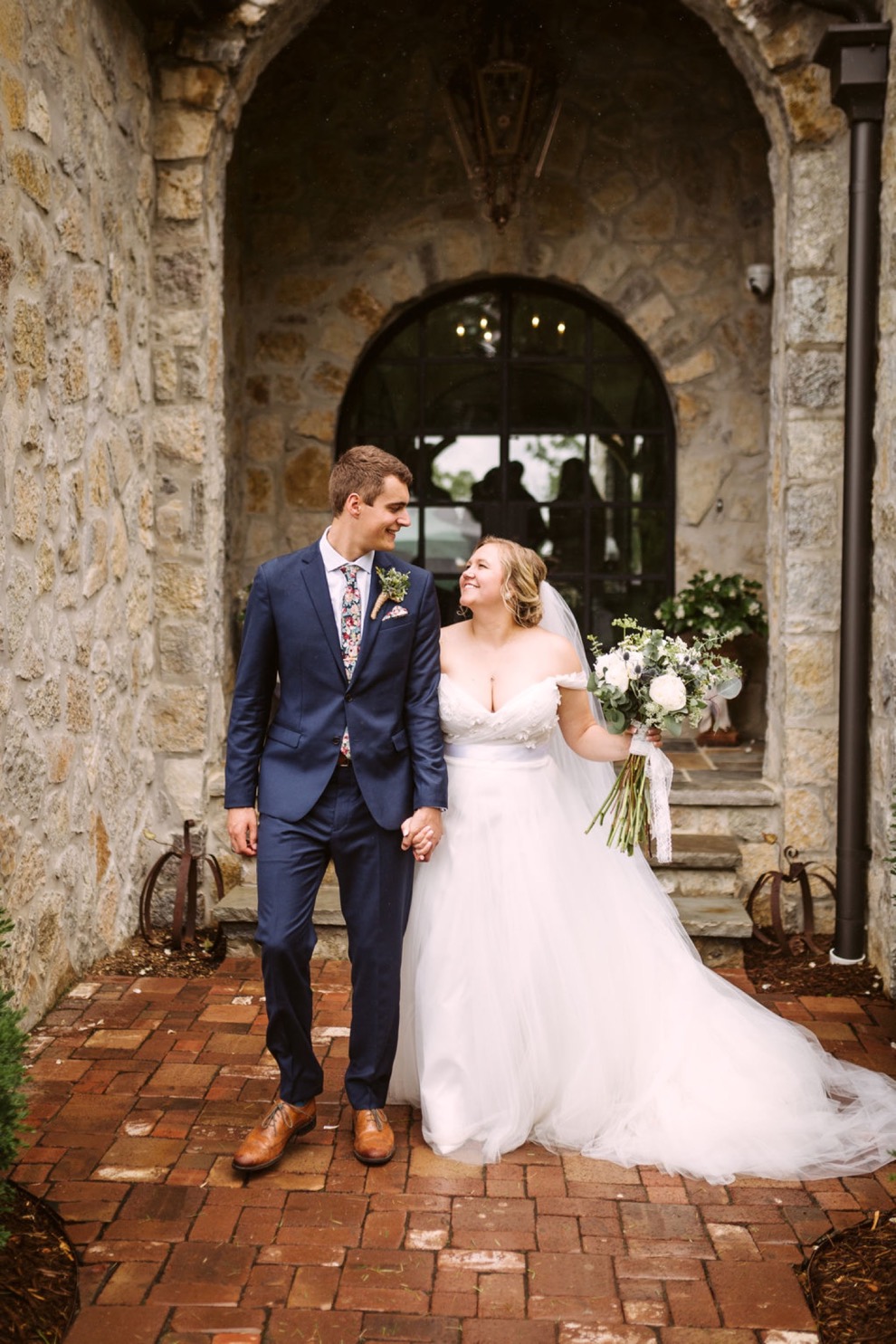 bride and groom walking hand in hand down a brick pathway in front of a stone house