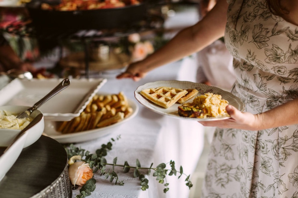 woman in floral printed dress adding eggs and waffles to her plate at backyard brunch wedding reception