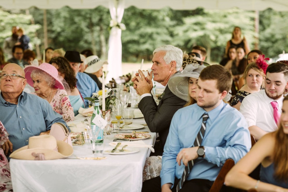 guests sitting at long tables under wedding tent listening to wedding toasts