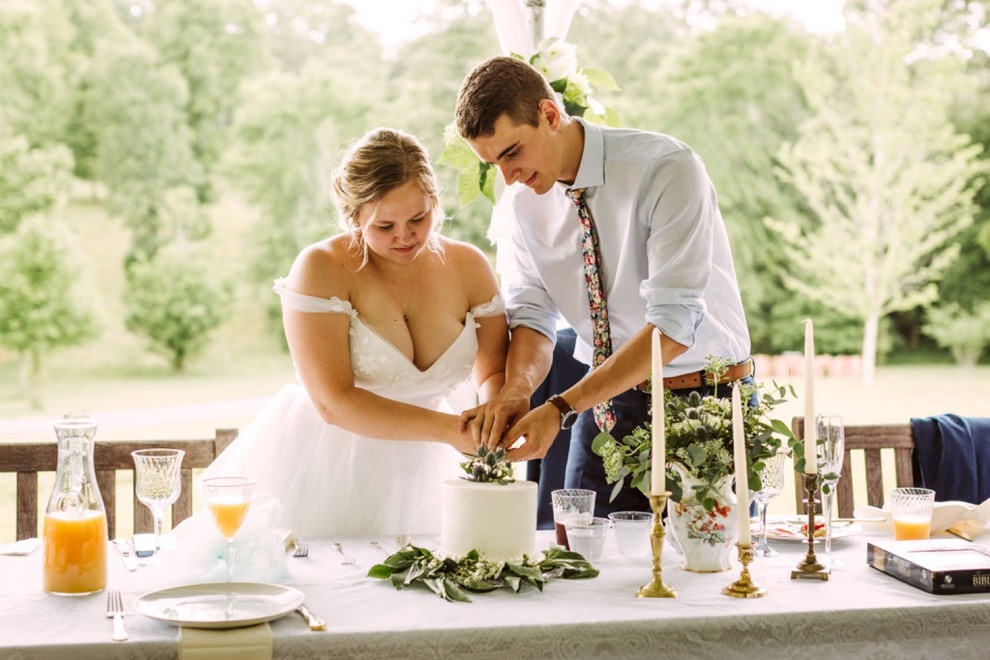 bride and groom cutting wedding cake. A glass carafe of orange juice sits on the table next to them.